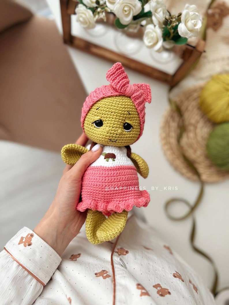 May include: A crocheted doll with a pink dress and headband. The doll has a yellow body and a white shirt with a brown turtle design. The doll is being held by a hand.