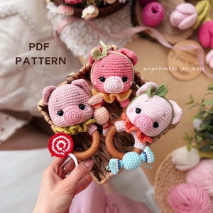 以下が含まれることがあります： 編みぐるみの豚の形をしたベビーラトル3つが、柳のバスケットに入っています。各豚はピンク色の体、木のリング、カラフルな襟が付いています。1つのラトルには赤と白のロリポップ、別のラトルには青いキャンディー、3つ目には緑色の襟が付いています。