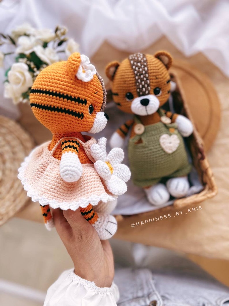 May include: Two crocheted tiger plushies. One is wearing a pink dress with a white flower and the other is wearing a green overall with a heart. The plushies are sitting on a wooden surface.