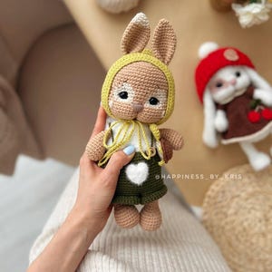 May include: A crocheted bunny toy wearing a yellow hat and green dungarees with a white heart. The bunny has brown fur and white accents on its face.