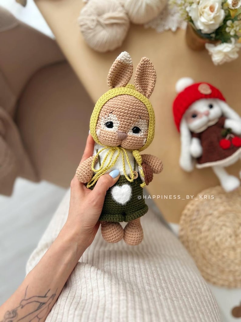 May include: A crocheted bunny toy wearing a yellow hat and green dungarees with a white heart. The bunny has brown fur and white accents on its face.