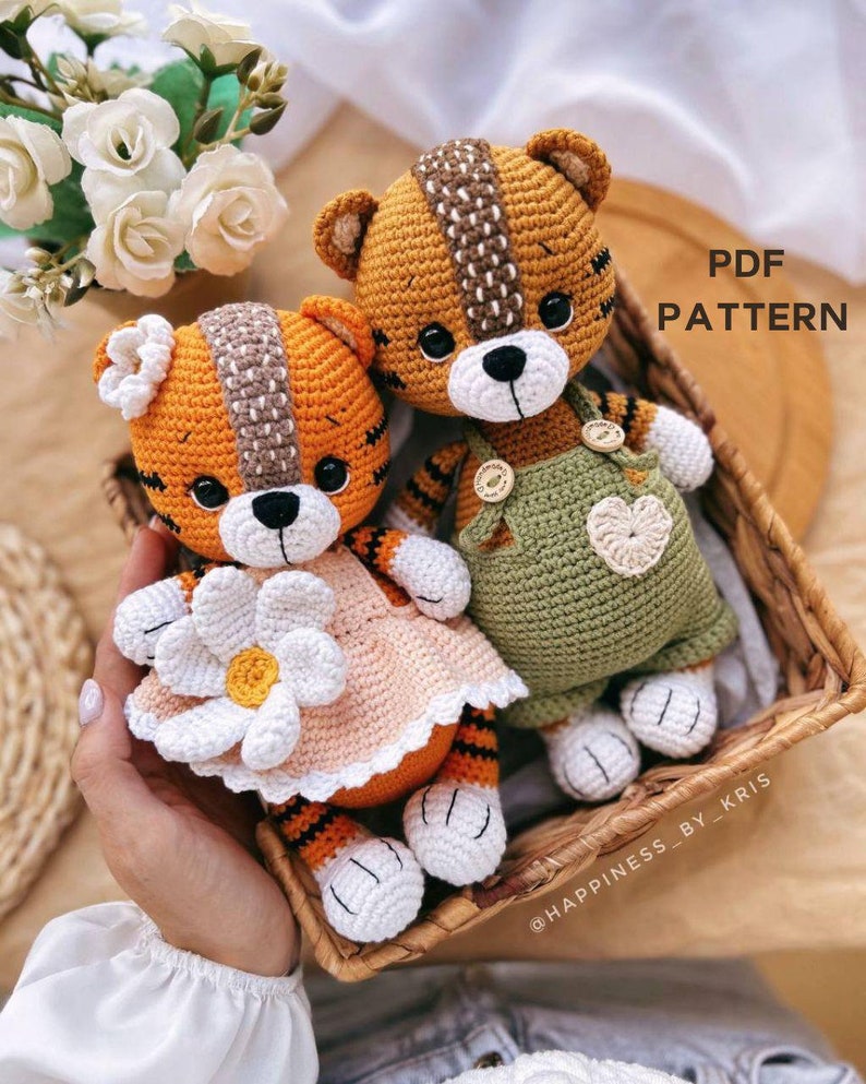 May include: Two crocheted tiger plushies, one wearing a peach dress with a white flower and the other wearing green overalls with a heart. The plushies are sitting in a wicker basket. The text "PDF PATTERN" is in the upper right corner of the image.