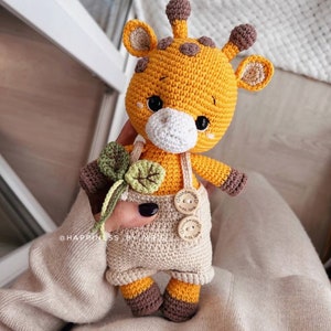 May include: A crocheted giraffe doll with a yellow body, brown spots, and brown accents. The giraffe wears beige overalls with wooden buttons. It holds a green and beige crocheted flower. The doll has black eyes and a white muzzle.