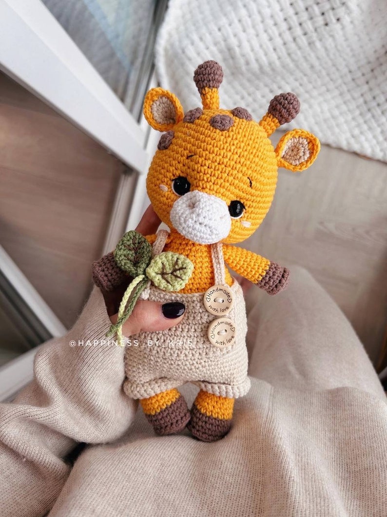 May include: A crocheted giraffe doll with a yellow body, brown spots, and brown accents. The giraffe wears beige overalls with wooden buttons. It holds a green and beige crocheted flower. The doll has black eyes and a white muzzle.