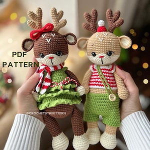 May include: Two crocheted reindeer toys, one wearing a green dress with a red and white striped scarf and the other wearing a red and white striped shirt and green shorts. Both reindeer have brown antlers and are holding a red bow.