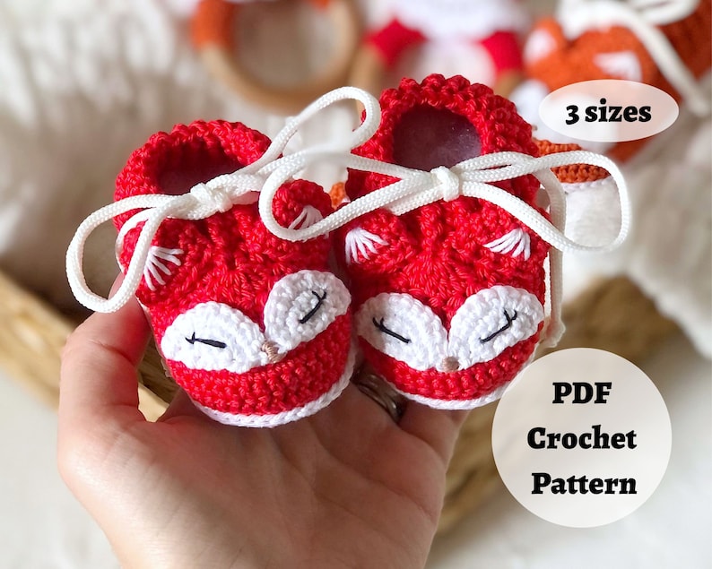 May include: Red crocheted baby booties with white fox faces and white laces. The booties are in a hand. The text "3 sizes" and "PDF Crochet Pattern" is on the image.