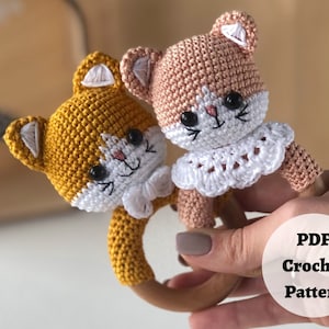 May include: Two crocheted cat rattle toys with wooden rings. One cat is yellow and the other is pink. Both cats have white faces with black eyes and whiskers.