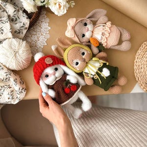 May include: Three crocheted bunny plushies with different coloured outfits. The bunny on the left is wearing a red hat and a brown dress with cherries. The bunny in the middle is wearing a green and yellow outfit. The bunny on the right is wearing a pink dress with a bow.