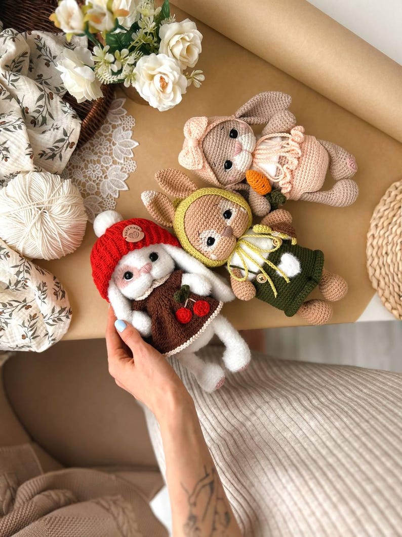 May include: Three crocheted bunny plushies with different coloured outfits. The bunny on the left is wearing a red hat and a brown dress with cherries. The bunny in the middle is wearing a green and yellow outfit. The bunny on the right is wearing a pink dress with a bow.