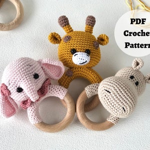May include: Three crocheted animal toys with wooden rings. The toys are a pink elephant, a yellow giraffe, and a beige hippopotamus. The toys are all handmade and have a cute, whimsical look. PDF Crochet Pattern.