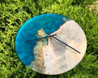 May include: A round wall clock with a blue resin and wood face. The clock has black hands and a gold center. The resin portion of the clock face resembles a body of water with a wood shoreline.