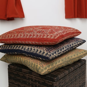 May include: Three patterned throw cushions stacked on top of a woven wicker storage bin. The top cushion is red with a geometric design, the middle cushion is navy blue with a geometric design, and the bottom cushion is green with a geometric design.