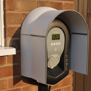 Pode incluir: Um carregador de veículo elétrico branco e cinza montado em uma parede de tijolos. O carregador possui um visor digital, uma porta de carregamento e o nome da marca "zappi". O carregador é parcialmente coberto por uma capa cinza.