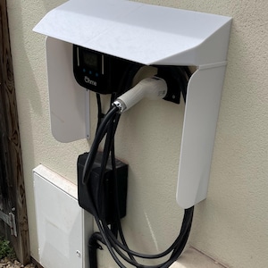 May include: A white protective cover mounted over an Ohme electric vehicle charging station. The unit features a black display screen, a black charging cable, and a white charging connector nozzle docked securely within the protective housing.