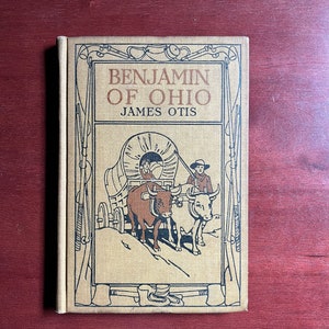 May include: A vintage book cover with a tan background and a black and white illustration of a covered wagon pulled by two oxen. The title of the book is "Benjamin of Ohio" by James Otis.