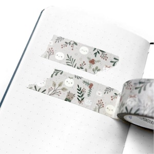 May include: A roll of washi tape with a white background and a repeating pattern of small white ghosts, green leaves, red berries, and brown presents. The tape is partially unrolled and there are two pieces of tape already placed on a white dotted journal page.