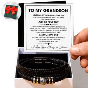 Puede incluir: Una pulsera de cuero negro con detalles plateados y dorados se muestra en una caja de regalo abierta. La caja contiene una tarjeta con el texto "TO MY GRANDSON" y un mensaje. Una pequeña caja de regalo verde con un lazo rojo está en la esquina superior izquierda.