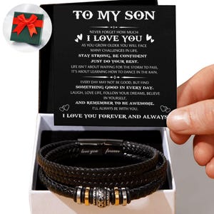 Peut inclure: Un bracelet en cuir noir avec des détails argentés et dorés, présenté dans un coffret cadeau blanc. La boîte contient une carte avec le message "To My Son" et un message affectueux. Un petit coffret cadeau vert avec un nœud rouge est également visible.