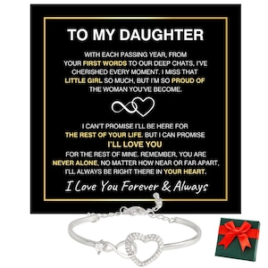 Puede incluir: Una pulsera plateada con un colgante en forma de corazón y un símbolo de infinito, junto a una tarjeta negra con ribete dorado y el texto "TO MY DAUGHTER". También hay una pequeña caja de regalo verde con un lazo rojo.