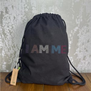 May include: Black drawstring bag featuring the text "I AM ME" in a dotted pattern. The letters are in shades of brown, white, blue, and red. The bag has black drawstrings and a small tag.