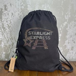May include: Black drawstring bag featuring the text "STARLIGHT EXPRESS" in a rhinestone design. The bag has a star graphic and a small tag attached. The bag is made of a soft material and has black drawstrings.