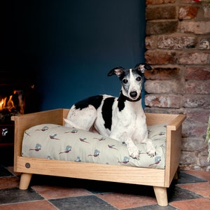 May include: A black and white dog lying in a wooden dog bed with a green and brown patterned cushion. The bed is in front of a fireplace with a wood-burning stove and a basket of logs.