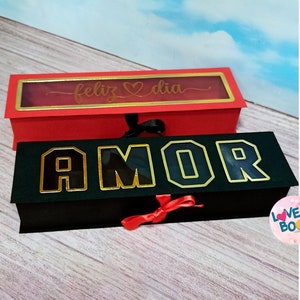 Puede incluir: Dos diseños para una caja de libro, una roja con una ventana y el texto "feliz dia" y otra negra con la palabra "AMOR" en letras doradas. Las cajas miden 9,8 pulgadas por 2,8 pulgadas.