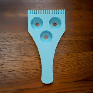 May include: A light blue plastic tool with three round holes and a comb-like top. The tool is used for making donuts.