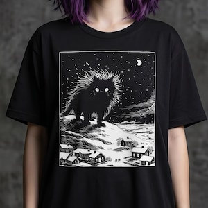 以下が含まれることがあります： 黒いTシャツで、夜の小さな村を見下ろす雪の丘の上に立つ、毛並みの良い大きな目の猫の白いグラフィックプリントが施されています。イラストには、木版画風の星空と三日月が描かれています。