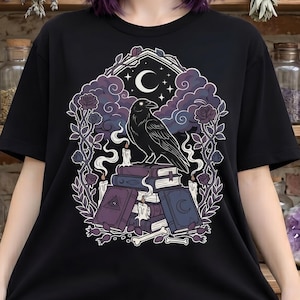 Pastel goth crow tshirt, featuring a crescent moon, spell books and candles. Unisex fit, crew neck and 100% cotton.