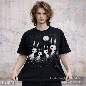 Pu&ograve; includere: T-shirt nero con un'illustrazione bianca di quattro creature simili a conigli sotto una luna piena. Il design include un cielo stellato e erba. Il marchio "KAMIOJI" &egrave; stampato in basso a sinistra. Il modello &egrave; alto 180 cm e indossa la taglia XL.