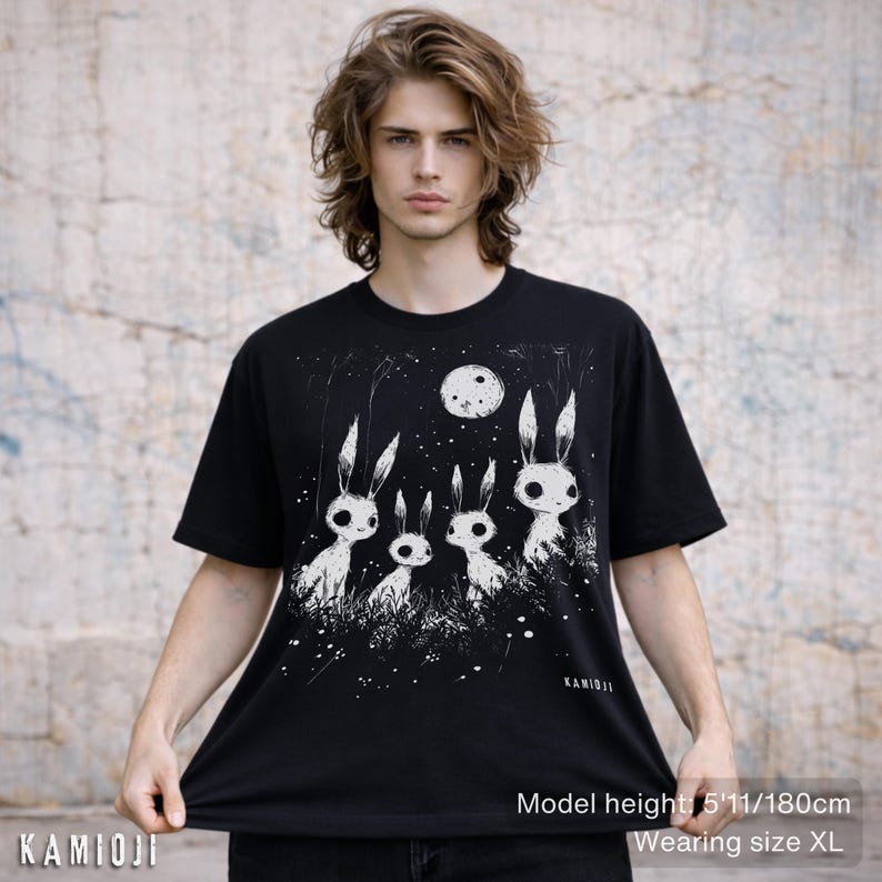Pu&ograve; includere: T-shirt nero con un'illustrazione bianca di quattro creature simili a conigli sotto una luna piena. Il design include un cielo stellato e erba. Il marchio "KAMIOJI" &egrave; stampato in basso a sinistra. Il modello &egrave; alto 180 cm e indossa la taglia XL.