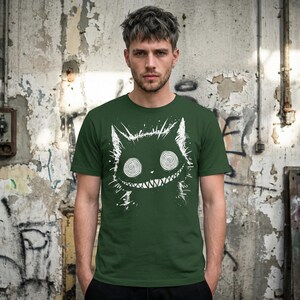 May include: Dark green t-shirt featuring a white, stylized cat graphic with spiral eyes and a toothy grin. The cat design has a distressed, hand-drawn aesthetic. The shirt is a crew neck and appears to be made of a soft material.