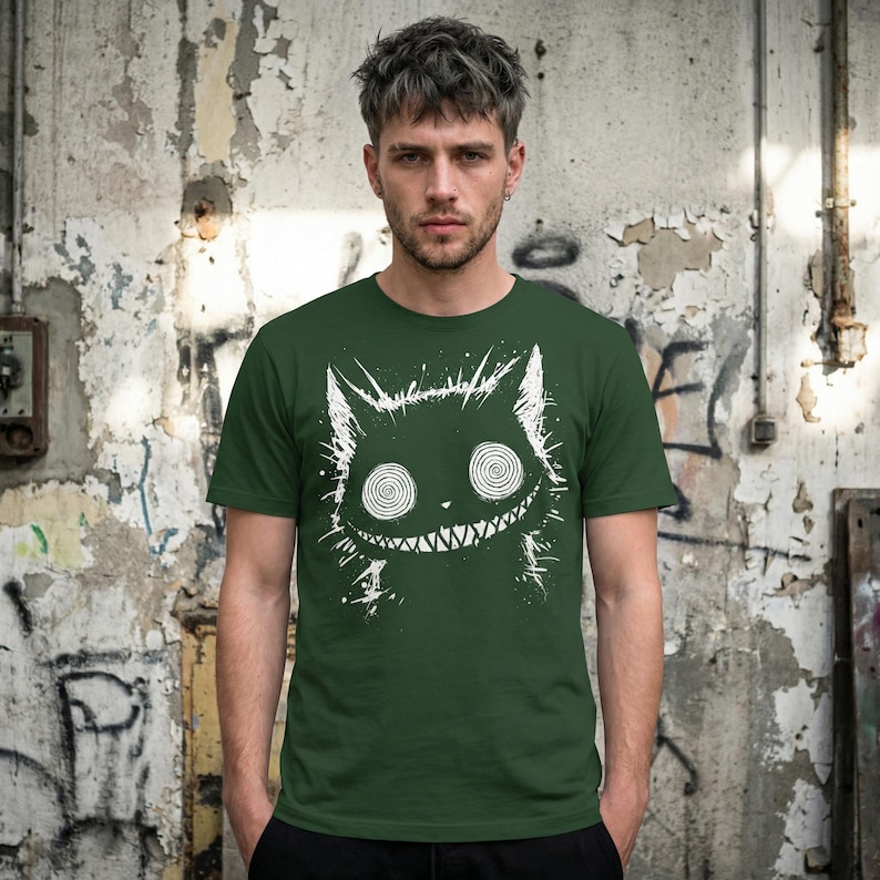 May include: Dark green t-shirt featuring a white, stylized cat graphic with spiral eyes and a toothy grin. The cat design has a distressed, hand-drawn aesthetic. The shirt is a crew neck and appears to be made of a soft material.