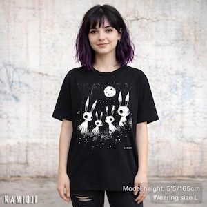 Pu&ograve; includere: T-shirt nero con grafica bianca di quattro conigli stilizzati sotto una luna piena. Il design include un cielo stellato e il testo "KAMIOJI". La modella indossa una taglia L. Altezza modella: 165 cm.