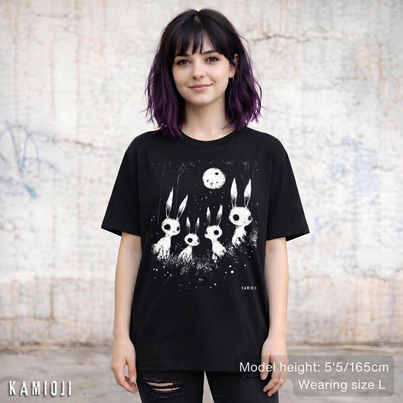 Pu&ograve; includere: T-shirt nero con grafica bianca di quattro conigli stilizzati sotto una luna piena. Il design include un cielo stellato e il testo "KAMIOJI". La modella indossa una taglia L. Altezza modella: 165 cm.