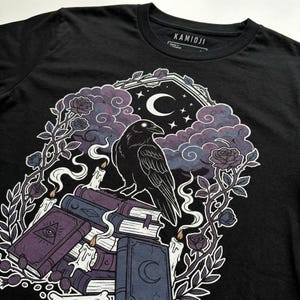 May include: Black t-shirt featuring a detailed illustration of a raven perched atop a stack of books, with a crescent moon, stars, and roses. The design includes candles and a mystical, gothic aesthetic. The brand name "KAMIOJI" is printed on the collar.