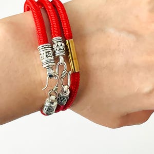 May include: A red string bracelet with silver and gold accents. The bracelet has three strands of red string and three silver charms. The charms are in the shape of a circle, a hook, and a bell. The bracelet is tied with a gold clasp.