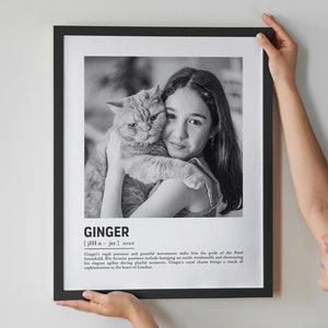 May include: Black and white photo of a girl hugging a cat. The photo is in a black frame and has a definition of the word "Ginger" below the photo. The definition reads: "Ginger's regal presence and graceful movements make him the pride of the Patel household. His favorite pastimes include lounging on sunlit windowsills and showcasing his elegant agility during playful moments. Ginger's royal charm brings a touch of sophistication to the heart of London."
