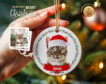 May include: A white ceramic Christmas ornament with a red and green wreath design. A cat wearing a Santa hat is in the center of the ornament. The cat's name, "Sandy", is written on a red ribbon below the cat.