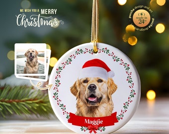 May include: A white ceramic Christmas ornament with a red and green wreath border. A golden retriever dog wearing a red Santa hat is in the center of the ornament. The dog's name, "Maggie", is written on a red ribbon.