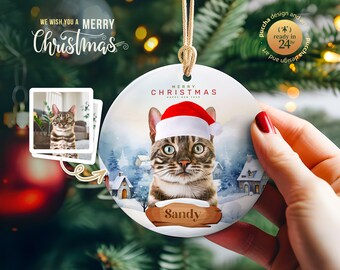 May include: A round, white ceramic ornament with a winter scene featuring a cat wearing a Santa hat. The cat is looking at the camera and has a name tag that says "Sandy". The ornament has the text "Merry Christmas Happy New Year" on it.