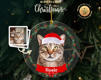 May include: A clear, round Christmas ornament featuring a tabby cat wearing a Santa hat. The ornament has a red banner with the name "Rosie" and a green wreath design. The text "We wish you a Merry Christmas" is at the top.