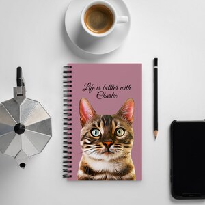 May include: A pink spiral bound notebook with a brown tabby cat illustration and the text "Life is better with Charlie".