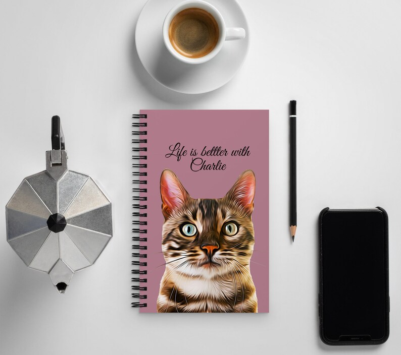 May include: A pink spiral bound notebook with a brown tabby cat illustration and the text "Life is better with Charlie".