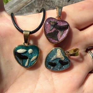 May include: Three heart-shaped pendants with gold-toned metal frames. Two pendants feature teal glitter resin with embedded shark teeth and small shells. One pendant features purple glitter resin with embedded shark teeth. One has a black cord.