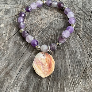 May include: A purple amethyst gemstone bracelet with a silver charm featuring a natural seashell. The bracelet is made with faceted beads and silver spacers.