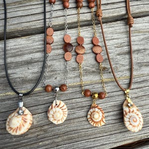 May include: Five necklaces with seashell pendants and beaded accents. The necklaces feature various chain and cord materials, including black cord, silver chain, gold chain, and brown leather cord. The pendants are cream-colored with brown spiral patterns.
