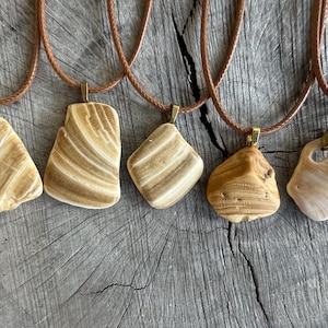 May include: Five handcrafted pendant necklaces with natural, beige-colored stone pendants. Each pendant is unique in shape and texture, suspended from a brown cord with a gold-colored clasp. The necklaces are displayed on a rustic, wooden surface.
