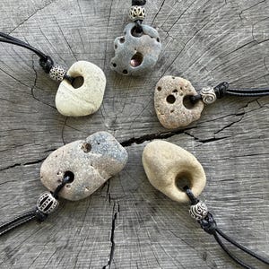 May include: Five pendants featuring naturally weathered stones with holes, each suspended on a black cord. The stones vary in color from light beige to grey and brown, and each is accented with a decorative silver-toned metal bead above the stone.